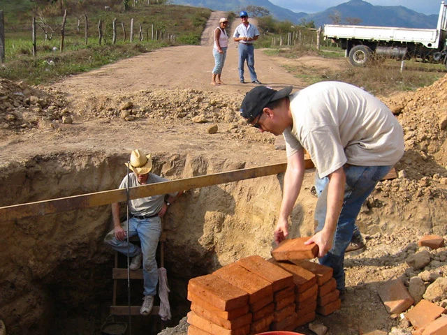 Man reaches over a pile of bricks to hand one to another volunteer who is standing at the bottom of a pit in the ground.