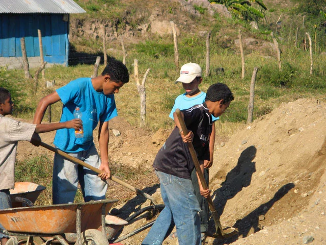 Three boys work at an outdoor construction site shoveling dirt into a wheelbarrow.