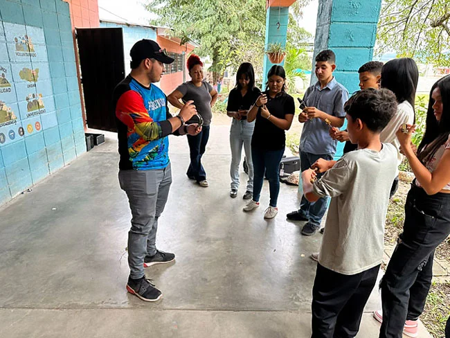 A group of band students holding instruments gather outside their school in a half circle to watch and listen to their music instructor.