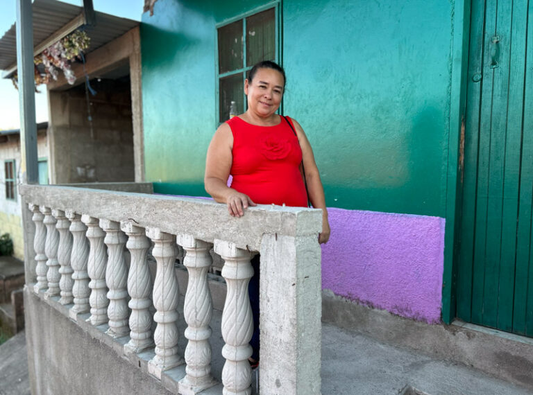 Woman smiles while standing outside on the front porch of her brightly painted teal and violet house in Colonia Solidaridad, Juticalpa.